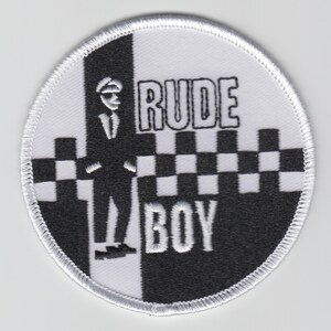 May include: A round patch with a white border featuring a black and white design. The patch is divided vertically, with a black and white checkerboard pattern on the right and a black silhouette of a man on the left. The words "RUDE BOY" are printed in black.