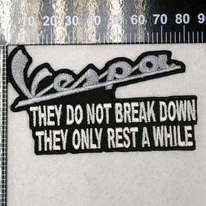 May include: A black and silver embroidered patch featuring the word Vespa in a script font above the text THEY DO NOT BREAK DOWN THEY ONLY REST A WHILE. The patch is positioned next to a ruler showing a width of approximately 3.5 inches.