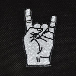 Könnte beinhalten: Ein weißer, gestickter Aufnäher in Form einer Hand, die das Rock'n'Roll-Zeichen zeigt. Der Aufnäher ist weiß umrandet und hat schwarze Details. Der Hintergrund ist ein strukturiertes schwarzes Gewebe.