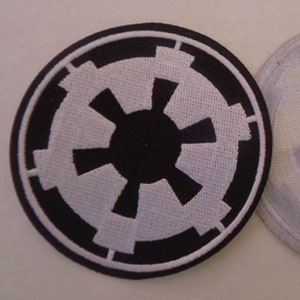 Puede incluir: Un parche bordado en blanco y negro con el símbolo imperial de Star Wars. El símbolo es un círculo con una estrella blanca en el interior, rodeado por un anillo negro.