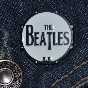 May include: A circular pin badge with the words "THE BEATLES" in black lettering on a white background. The badge is set against a denim fabric, with a metal button visible in the lower left corner.