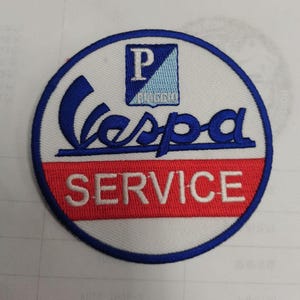 以下が含まれることがあります： 「Vespa SERVICE」の文字とピアジオのロゴが入った、白、青、赤の円形の刺繍パッチです。