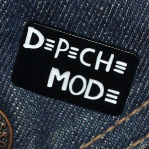 May include: Black enamel pin with white lettering that reads "D=PECH=MODE" on a denim background.