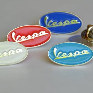 Op de afbeelding: Vier ovale metalen pinnen met het woord "Vespa" in witte letters op een blauwe, rode, witte en turquoise achtergrond.