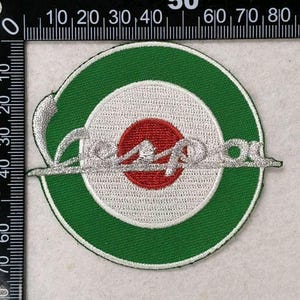 May include: A circular embroidered patch featuring a green outer ring, a white middle ring, and a red center circle. The silver-colored script text "Vespa" is embroidered across the center. A black ruler shows the patch is approximately 3.5 inches wide.