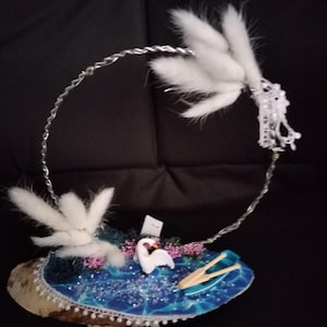 May include: A decorative swan-themed art piece. It features a wooden base with a blue, glittery water effect, a white swan, and a small boat with oars. White fluffy accents and a silver wire frame complete the design.