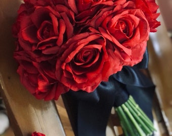 May include: A bouquet of red roses, tied with a black satin ribbon and green stems. A matching red rose buttonhole with a black stem is also present. The bouquet is on a wooden surface.