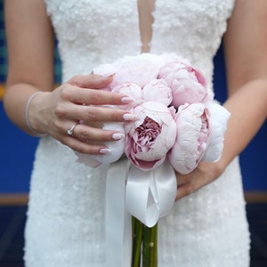 May include: A bride holds a bouquet of pink peonies with a white satin ribbon. The bouquet is held in front of a white wedding dress.