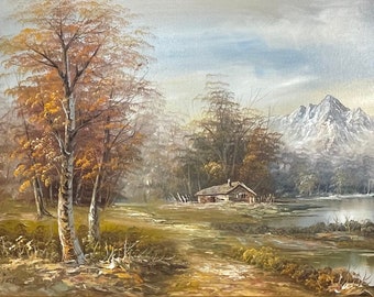 May include: A framed oil painting depicting a picturesque landscape with a small cabin nestled in a forest by a lake. The scene features a snow-capped mountain in the background and a winding river flowing through the foreground. The painting is framed in a gold-colored frame with ornate details.