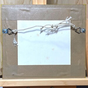 以下が含まれることがあります： 白いマットと茶色のフレームが付いた額縁の裏側。フレームには金属製の吊り下げフックが2つと、フックに白い紐が取り付けられています。