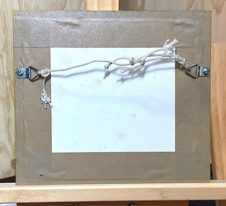 以下が含まれることがあります： 白いマットと茶色のフレームが付いた額縁の裏側。フレームには金属製の吊り下げフックが2つと、フックに白い紐が取り付けられています。