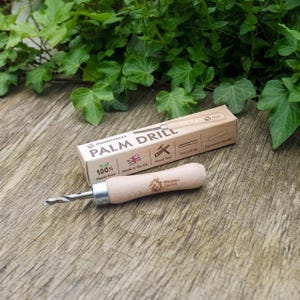 May include: A palm drill with a wooden handle and a silver drill bit, alongside its cardboard packaging. The packaging reads "PALM DRILL" and "Made in the UK". The drill is on a wooden surface with green foliage in the background.