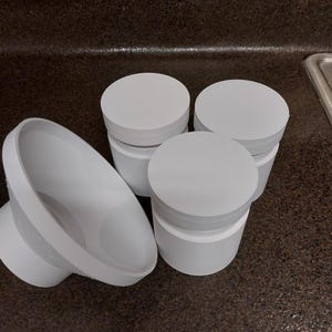 May include: Several white, cylindrical containers with lids are arranged on a dark countertop. One container is on its side, showing its interior. The containers are made of a solid white material.