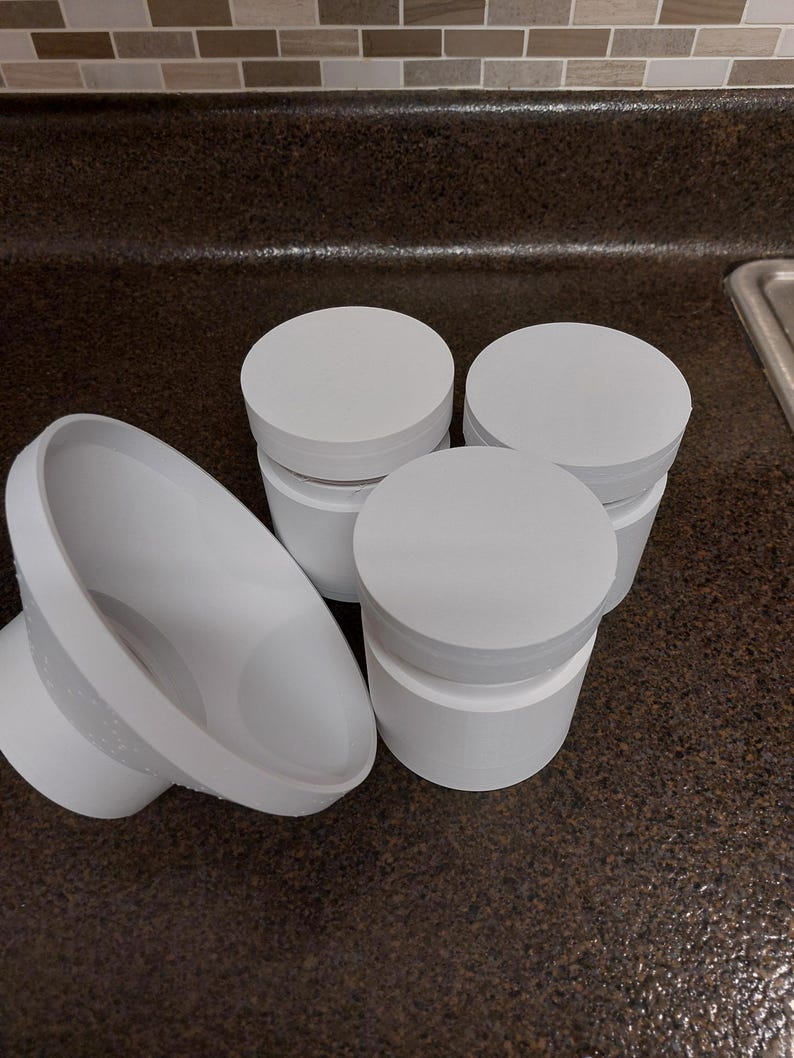 May include: Several white, cylindrical containers with lids are arranged on a dark countertop. One container is on its side, showing its interior. The containers are made of a solid white material.