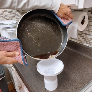 May include: A person pouring oil from a used frying pan into a white funnel. The pan is held with a pink and blue pot holder. The funnel is on a brown countertop. A roll of paper towels hangs on the wall.