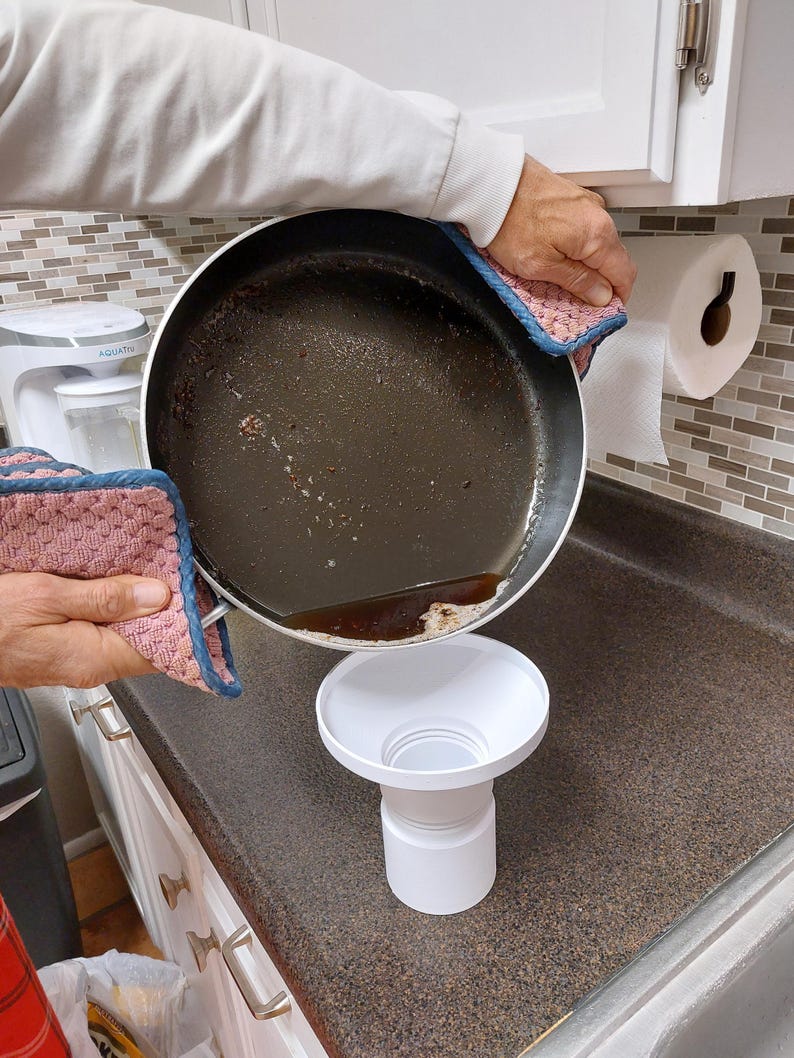 May include: A person pouring oil from a used frying pan into a white funnel. The pan is held with a pink and blue pot holder. The funnel is on a brown countertop. A roll of paper towels hangs on the wall.
