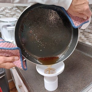 May include: A person pouring liquid from a dark pan into a white funnel. The pan is held with pink and blue oven gloves. The funnel is positioned over a white container on a brown countertop. The liquid is a golden brown colour.