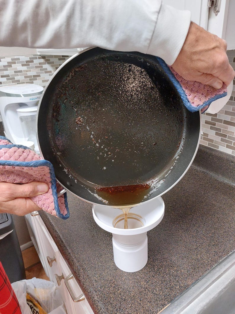 May include: A person pouring liquid from a dark pan into a white funnel. The pan is held with pink and blue oven gloves. The funnel is positioned over a white container on a brown countertop. The liquid is a golden brown colour.
