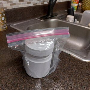 May include: A stack of white, cylindrical objects inside a clear plastic bag with a pink zip closure. The objects appear to be made of paper or a similar material. The bag is on a dark brown countertop near a kitchen sink.