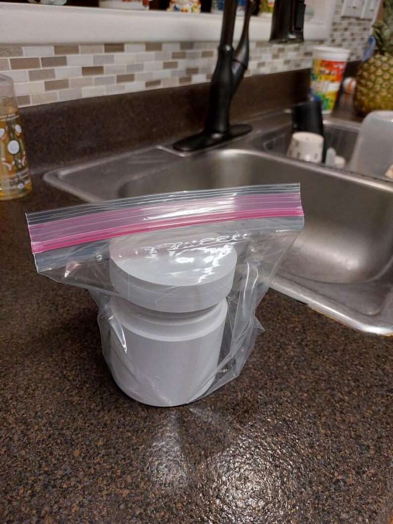 May include: A stack of white, cylindrical objects inside a clear plastic bag with a pink zip closure. The objects appear to be made of paper or a similar material. The bag is on a dark brown countertop near a kitchen sink.