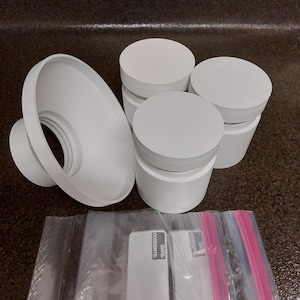 May include: A collection of white plastic containers with lids, a funnel-shaped object, and several clear plastic bags with pink and blue zip closures. The bags have white labels with the text "DATE: CONTENTS:" printed on them.