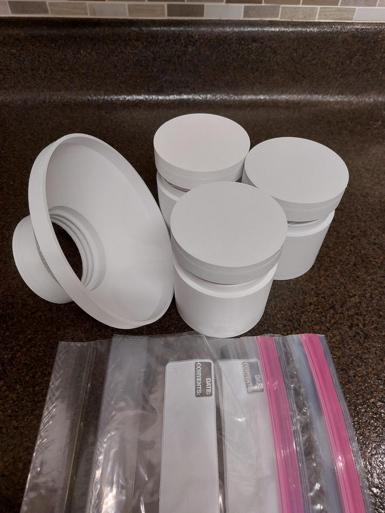 May include: A collection of white plastic containers with lids, a funnel-shaped object, and several clear plastic bags with pink and blue zip closures. The bags have white labels with the text "DATE: CONTENTS:" printed on them.