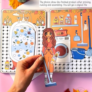 Pode incluir: Um conjunto de bonecas de papel imprim&iacute;veis com uma menina de cabelo comprido e ruivo que usa um su&eacute;ter vermelho e cal&ccedil;as laranja. A boneca vem com uma vassoura, um frasco de spray e um balde. O fundo mostra um banheiro com uma banheira, uma prateleira com produtos de limpeza e uma m&aacute;quina de lavar.