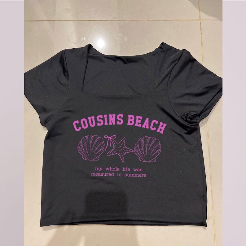 May include: Black square-neck crop top with short sleeves. The front features the text "COUSINS BEACH" in pink, along with seashell and starfish graphics. Below the graphics is the text "my whole life was measured in summers".