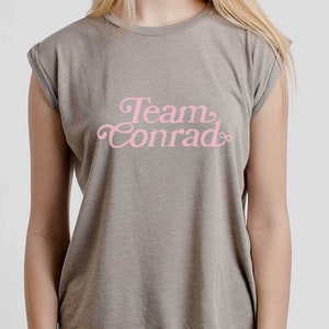 Op de afbeelding: Een grijs T-shirt met een roze afbeelding met de tekst "Team Conrad".