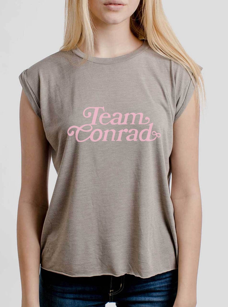 Op de afbeelding: Een grijs T-shirt met een roze afbeelding met de tekst "Team Conrad".