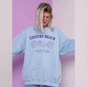 May include: Light blue sweatshirt with the text "COUSINS BEACH" above seashell and starfish graphics. Below the graphics is the text "my whole life was measured in summers". The sweatshirt is a casual, relaxed fit.