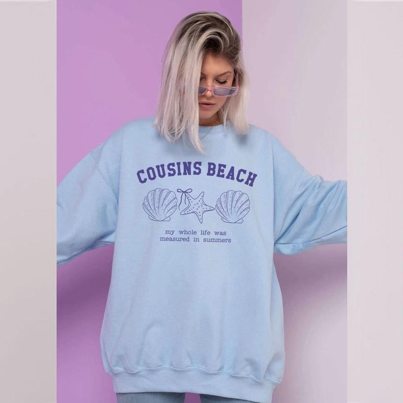 May include: Light blue sweatshirt with the text "COUSINS BEACH" above seashell and starfish graphics. Below the graphics is the text "my whole life was measured in summers". The sweatshirt is a casual, relaxed fit.