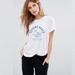 May include: White t-shirt with a blue graphic that says "COUSINS BEACH" in a circle with a sun and waves. Below the graphic is the text "my whole life was measured in summers". The shirt is being worn.