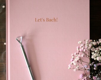 May include: A pink hardcover guest book with the text "Let's Bach!" printed in brown on the front cover. A silver pen with a diamond-shaped top is lying on the book. A bouquet of small pink and white flowers is on the right side of the book.