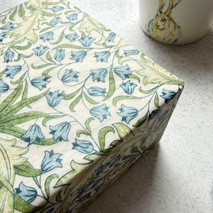 Pode incluir: Uma caixa decorativa com um padrão floral em tons de verde e azul. A caixa é adornada com um desenho repetido de flores em forma de sino e folhas. Uma caneca de cerâmica com uma ilustração de coelho e uma pequena garrafa de vidro também são visíveis.