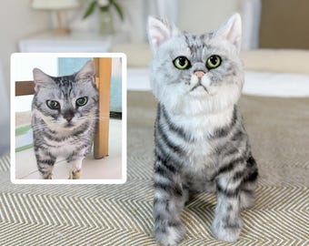 May include: A gray and white tabby cat plush toy sitting on a brown and white striped rug. The plush toy has a realistic appearance with a fluffy texture and detailed facial features.