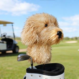 Puede incluir: Una funda para la cabeza del palo de golf con forma de perro golden retriever, con una textura de pelaje marrón claro y esponjoso. La funda está en una bolsa de golf con un palo de golf. Un carrito de golf está en el fondo en un campo de golf verde.