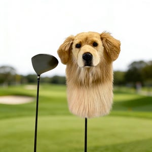 Könnte beinhalten: Eine Golfschlägerhaube in Form eines Golden Retriever Hundes. Die Haube ist beige und braun mit einem realistischen Hundegesicht und Fellstruktur. Ein schwarzer Golfschläger befindet sich neben der Haube auf einem grünen Golfplatz.