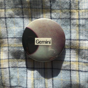 May include: A circular badge with a purple and grey design, including a dark crescent shape. The word "Gemini" is printed on a white rectangle. The badge is set against a blue and white checked fabric.