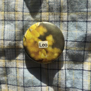 May include: A circular badge featuring a blurred image of yellow flowers and the word "Leo" in a white box. The badge is set against a blue and white checked fabric background.