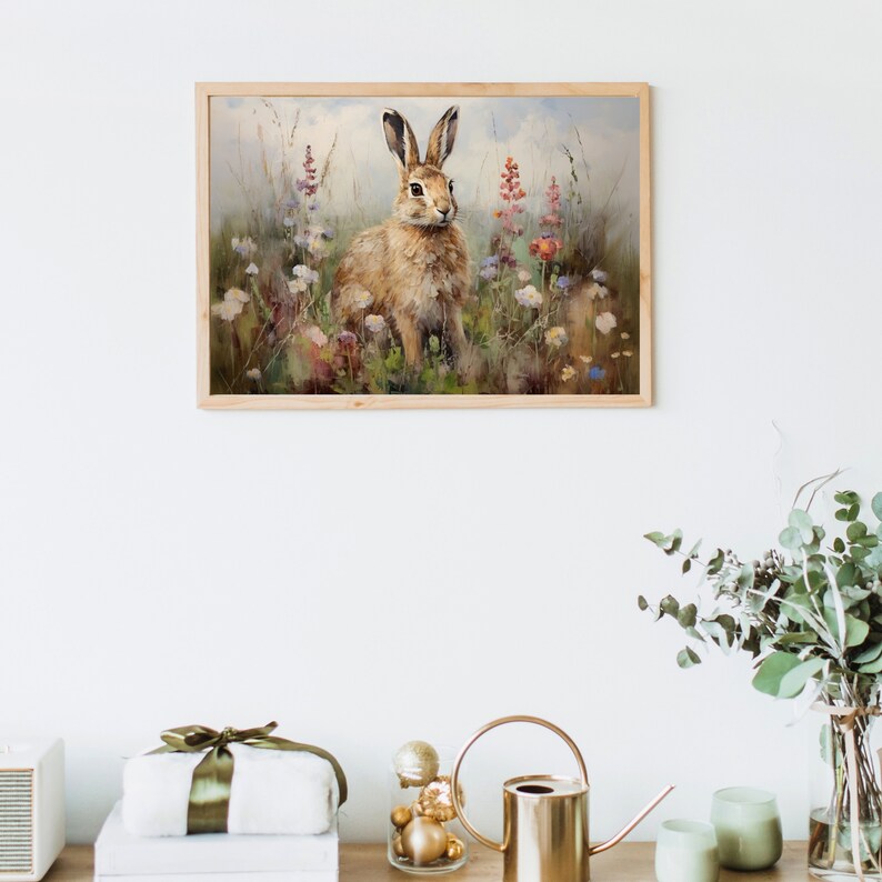 Może przedstawiać: Namalowany obraz w ramie przedstawiający brązowego kr&oacute;lika siedzącego na polu dzikich kwiat&oacute;w. Kr&oacute;lik patrzy bezpośrednio na widza. Tło jest miękkie, rozmyte w stylu akwareli.