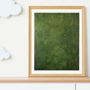 May include: A framed abstract art print with a textured, dark green background. The artwork is displayed in a light brown wooden frame with a white mat. The print is placed on a light brown wooden shelf, with white clouds on the left.