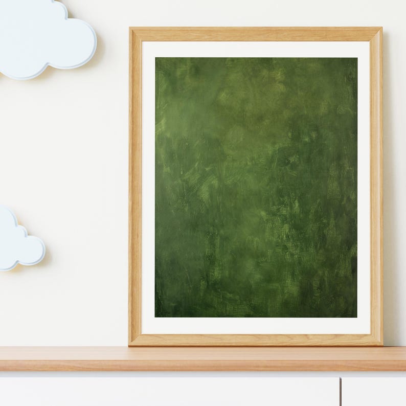 May include: A framed abstract art print with a textured, dark green background. The artwork is displayed in a light brown wooden frame with a white mat. The print is placed on a light brown wooden shelf, with white clouds on the left.