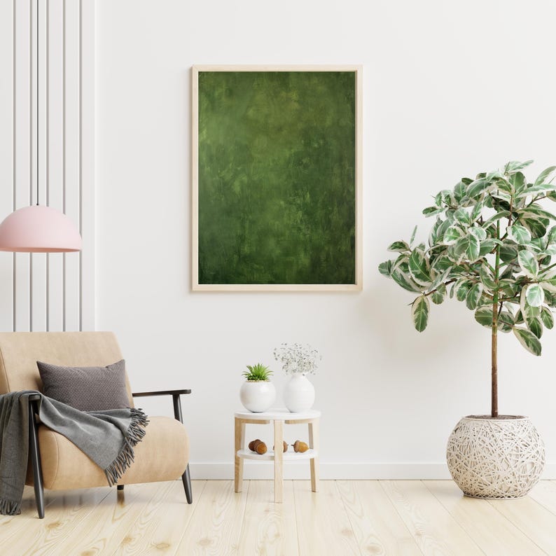 May include: A framed abstract painting with a textured green surface hangs on a white wall. The painting is in a light wooden frame. A beige armchair with a gray pillow and blanket sits below the painting. A potted plant and a side table are also in the room.