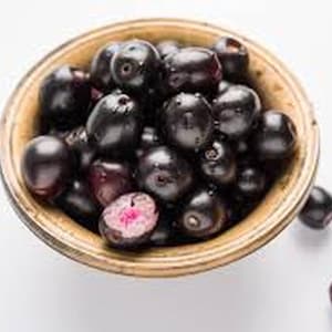 May include: A wooden bowl filled with dark purple java plums, also known as jambul or jamun. One plum is cut open, revealing a pink interior. The plums are on a white surface.