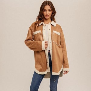 May include: A woman wearing a tan suede-like jacket with cream-colored trim and pockets, paired with blue jeans and cream-colored ankle boots. The jacket has a button-up front and a soft, textured appearance, suitable for fall or winter wear.
