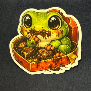 May include: A sticker featuring a cartoon frog with large eyes, covered in chocolate, inside a heart-shaped box of chocolates. The frog is green with brown accents, and the box is red with a gold ribbon.