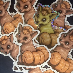Könnte beinhalten: Eine Sammlung von Aufklebern mit einem cartoonartigen, fuchsähnlichen Wesen mit einem kürbisförmigen Körper. Die Aufkleber sind hauptsächlich braun und orange, mit einem holografischen Aufkleber in der Mitte. Das Wesen hat große Augen und einen flauschigen Haarschopf.