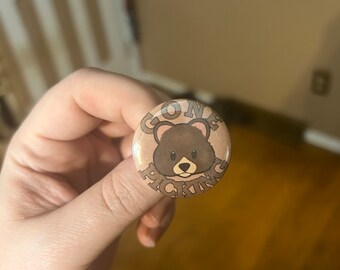 May include: A circular badge featuring a brown bear illustration and the text "GONE PICKING". The bear has a friendly face. The badge has a light brown background and is held in a hand.