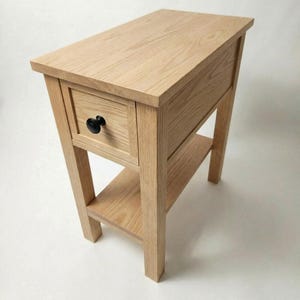 May include: A light-colored wooden side table featuring a rectangular top, a single drawer with a round black knob, and an open lower shelf. The table is constructed with four straight legs and displays a visible natural wood grain pattern throughout.